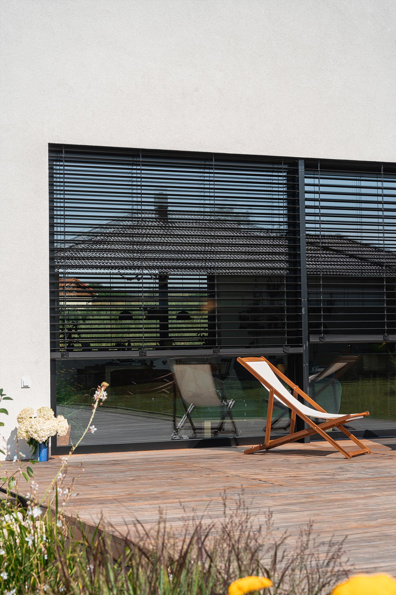 Large glass doors with dark horizontal blinds on a light stucco wall, reflecting a tiled roof and outdoor seating on a wooden deck
