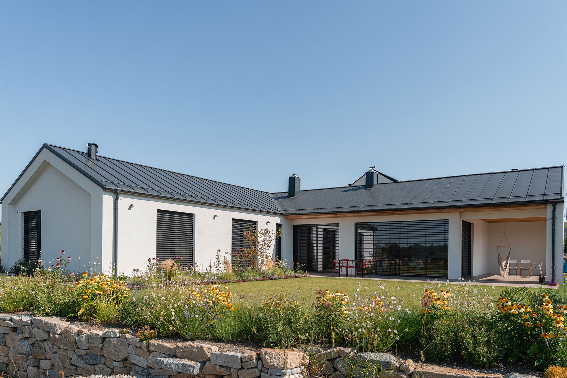 Modern single-story house with a dark metal roof, large windows, and a colorful flower garden in the foreground.