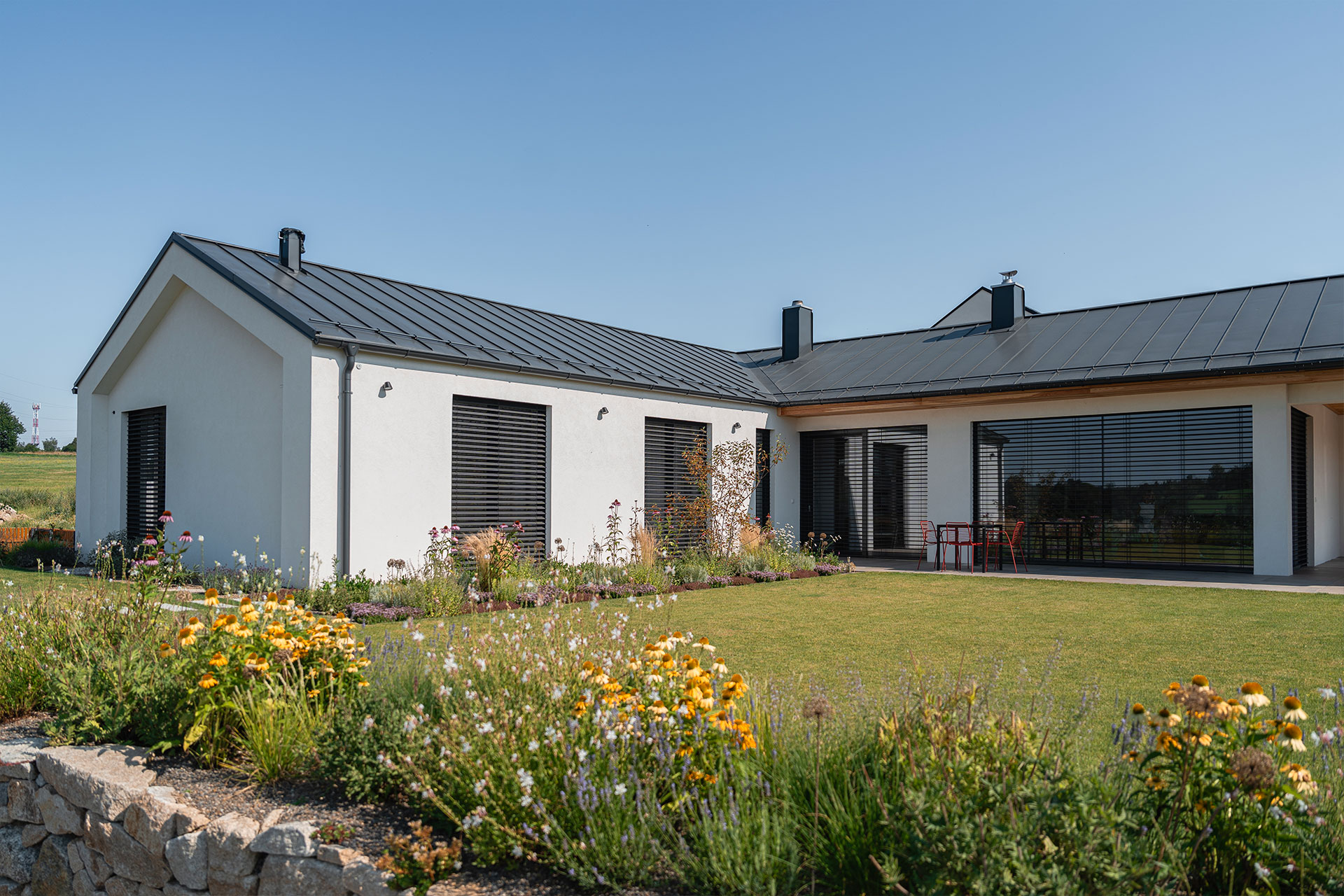 Modern single-story white house with a dark metal roof, large windows, and a landscaped flower garden in the foreground.]