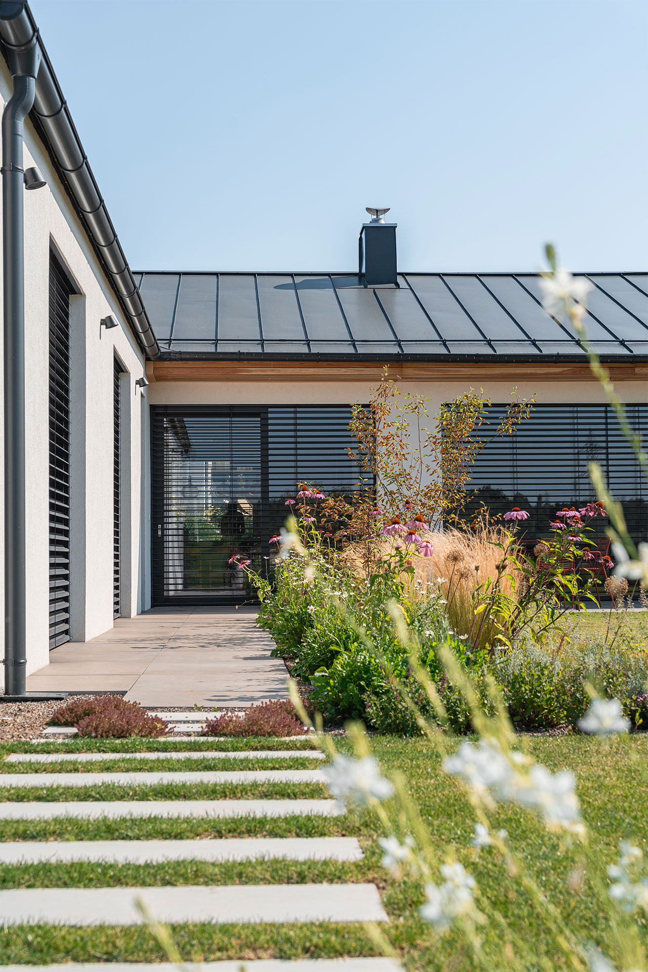 Modern house exterior with a metal roof, chimney and large glass doors/windows flanked by shutters, front garden in bloom.