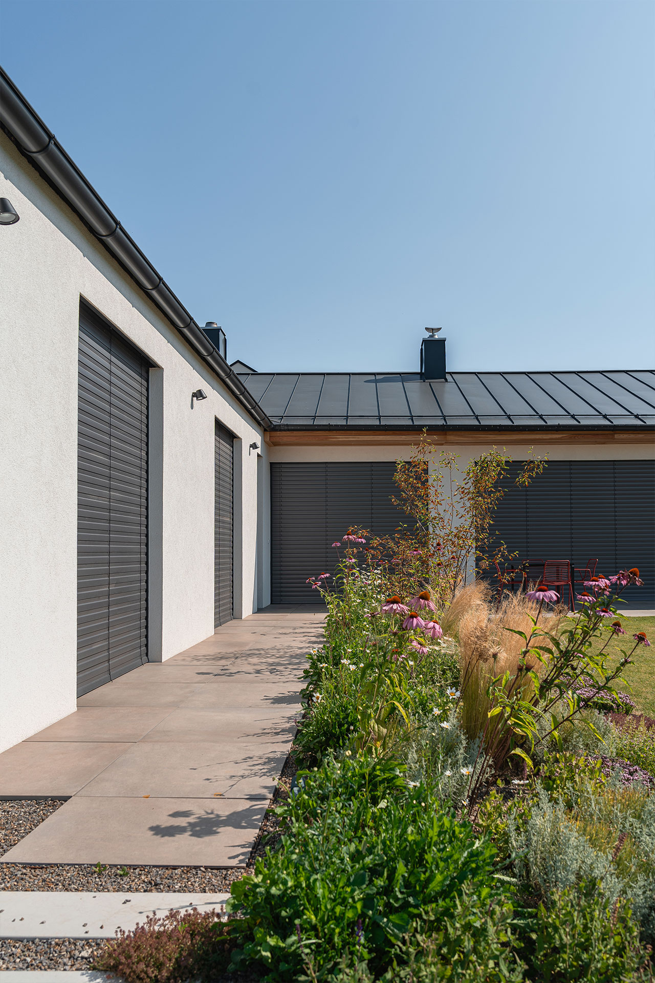 Modern house exterior with white stucco walls, dark gray roller shutters, and a landscaped garden along a paved path.