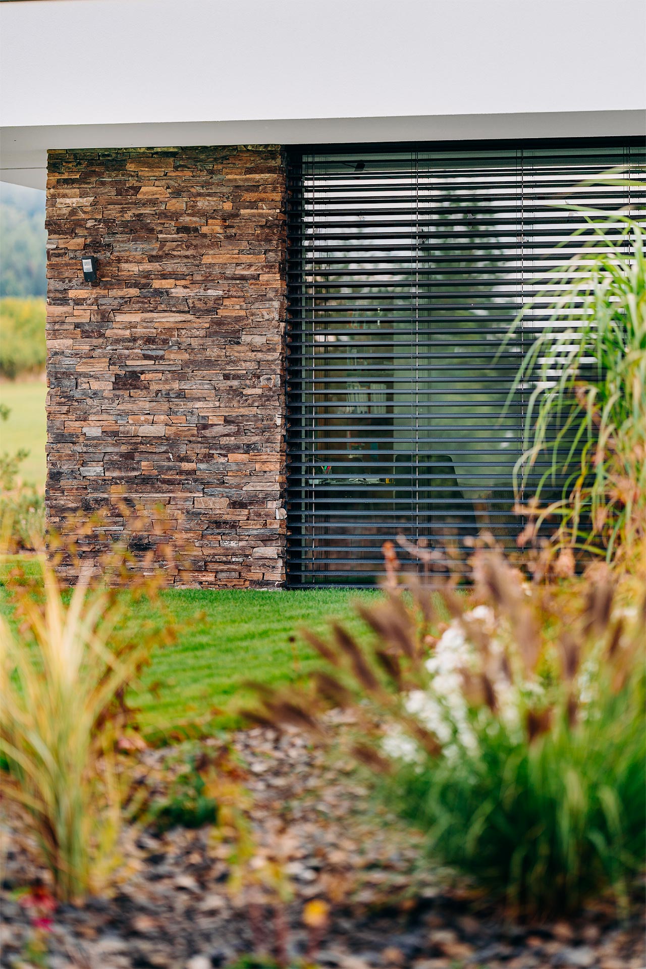 Modern house exterior with a stone façade pillar and a large window with horizontal blinds, framed by surrounding greenery.
