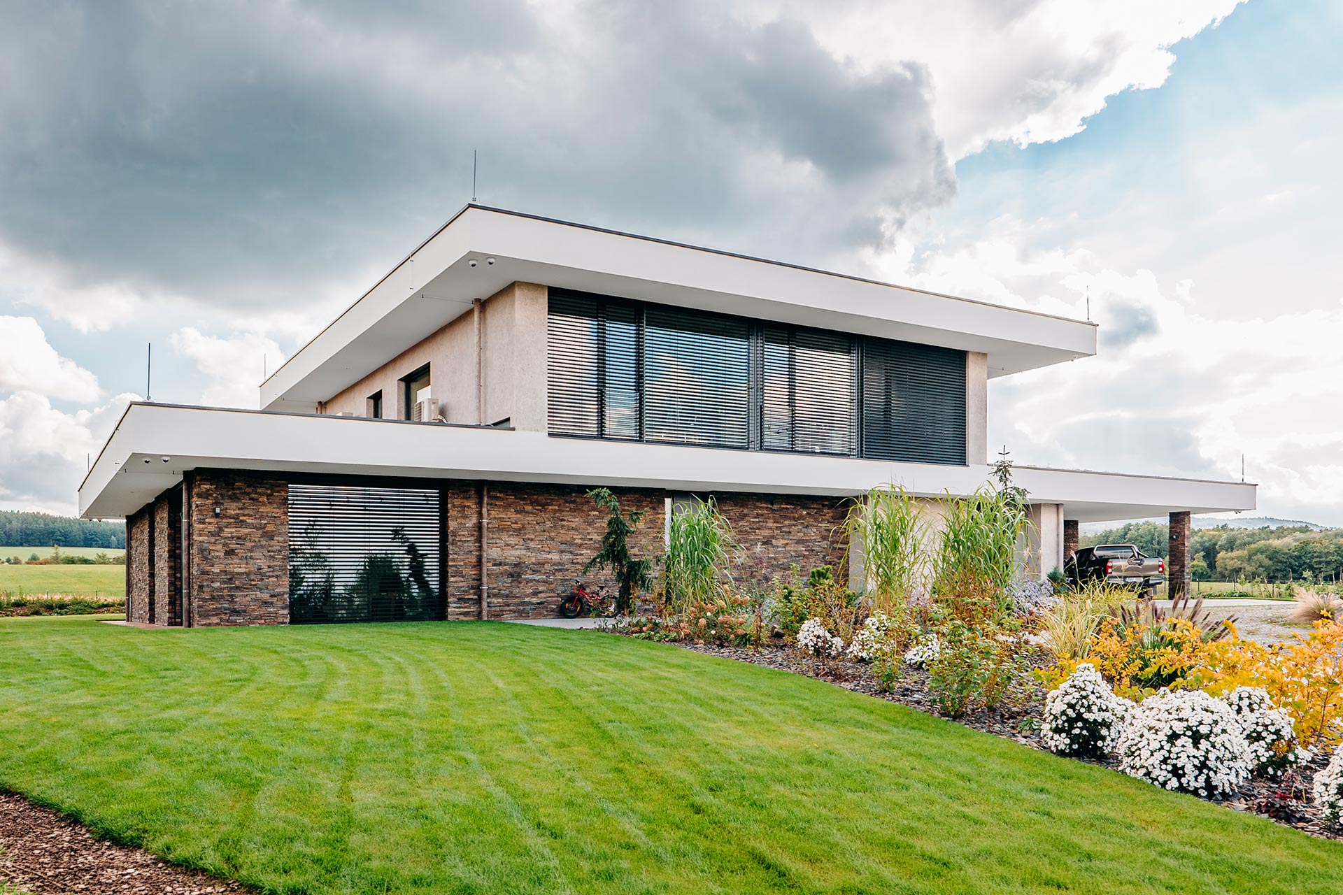 Modern two-story house with stone base, large glass windows with blinds, and manicured lawn in foreground.
