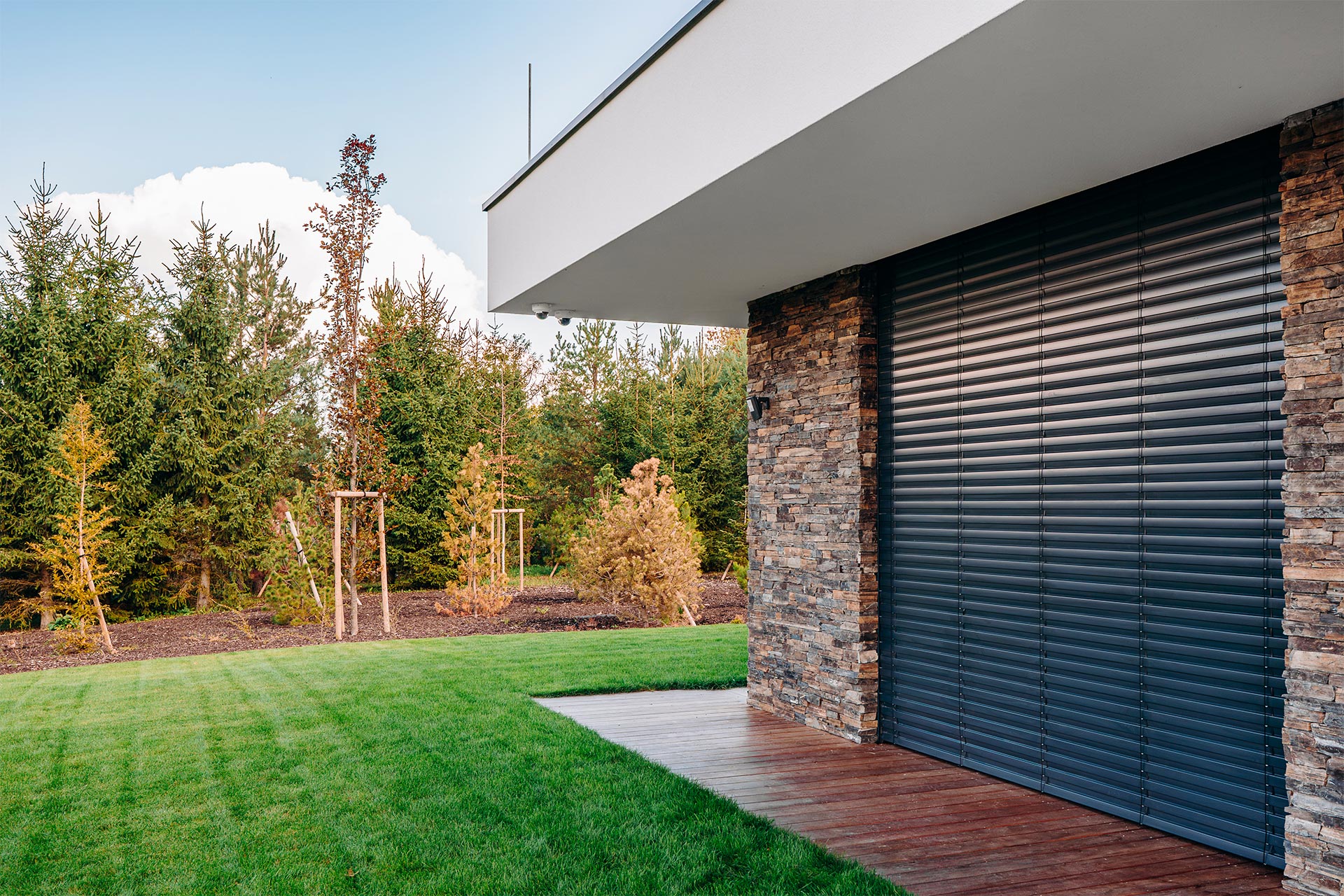 Modern house exterior with stone veneer, a large black roller shutter, and a wooden deck over a green lawn and blue sky.