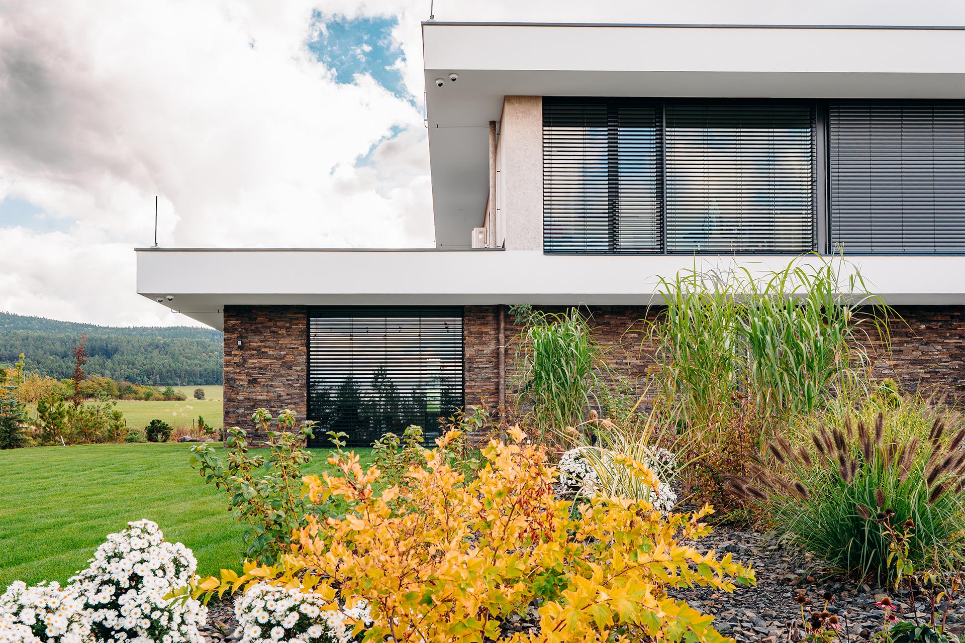 Modern two-story house with large windows and a stone ground floor, framed by autumn shrubs and a green lawn in the foreground.