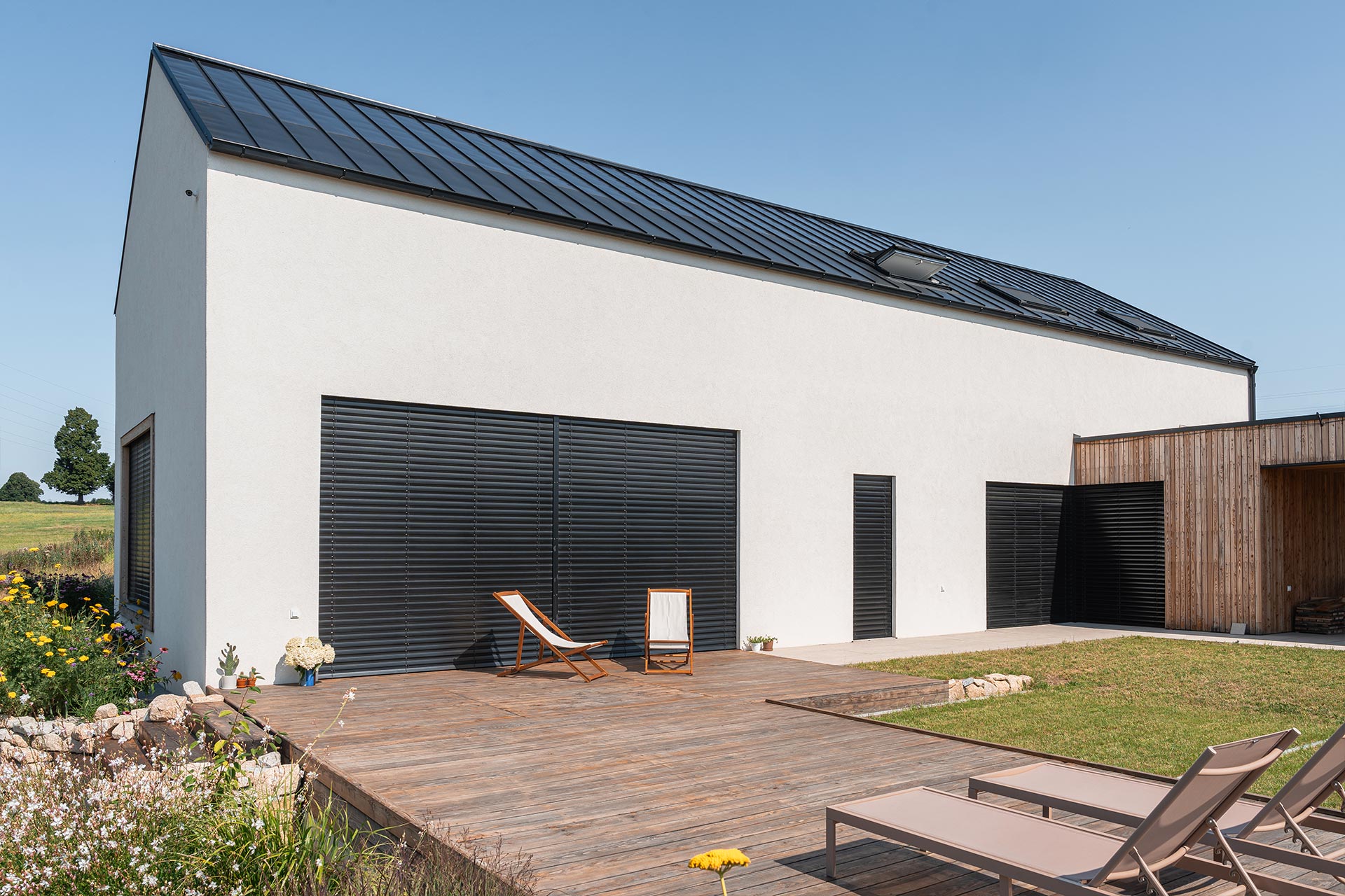 Modern white house with a black metal roof, large horizontal shutters, and a wooden deck with lounge chairs outside