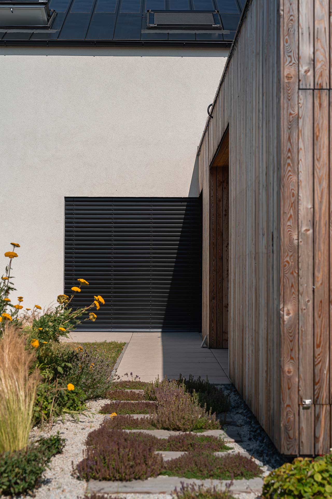 Modern building exterior with a white stucco wall, black horizontal siding, and a tall wooden fence on the right; a gravel path and yellow flowers in the foreground