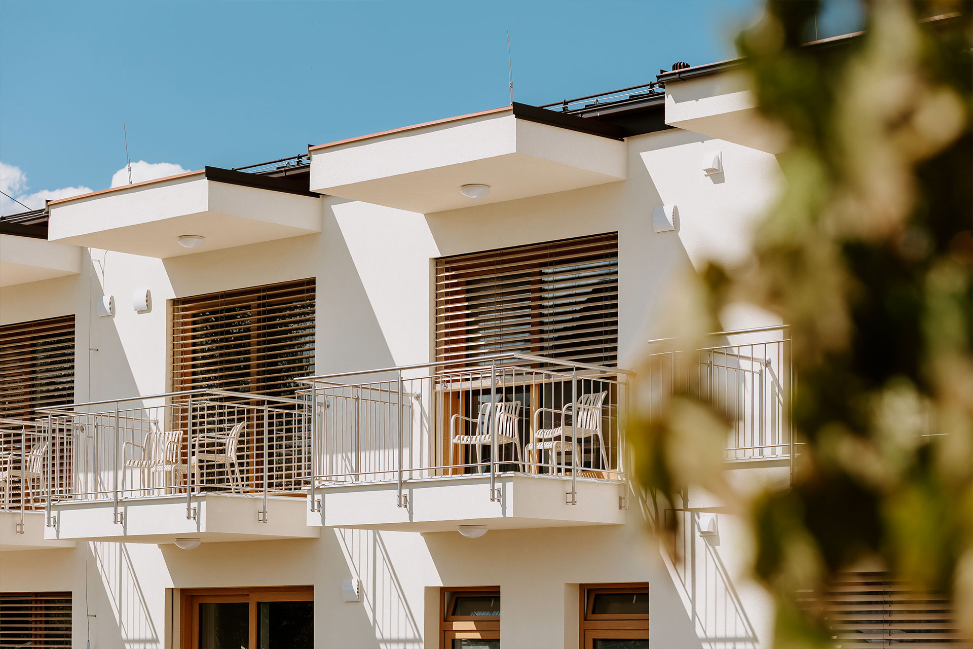 White apartment building with metal balcony railings, wooden window blinds, and outdoor chairs under a blue sky.