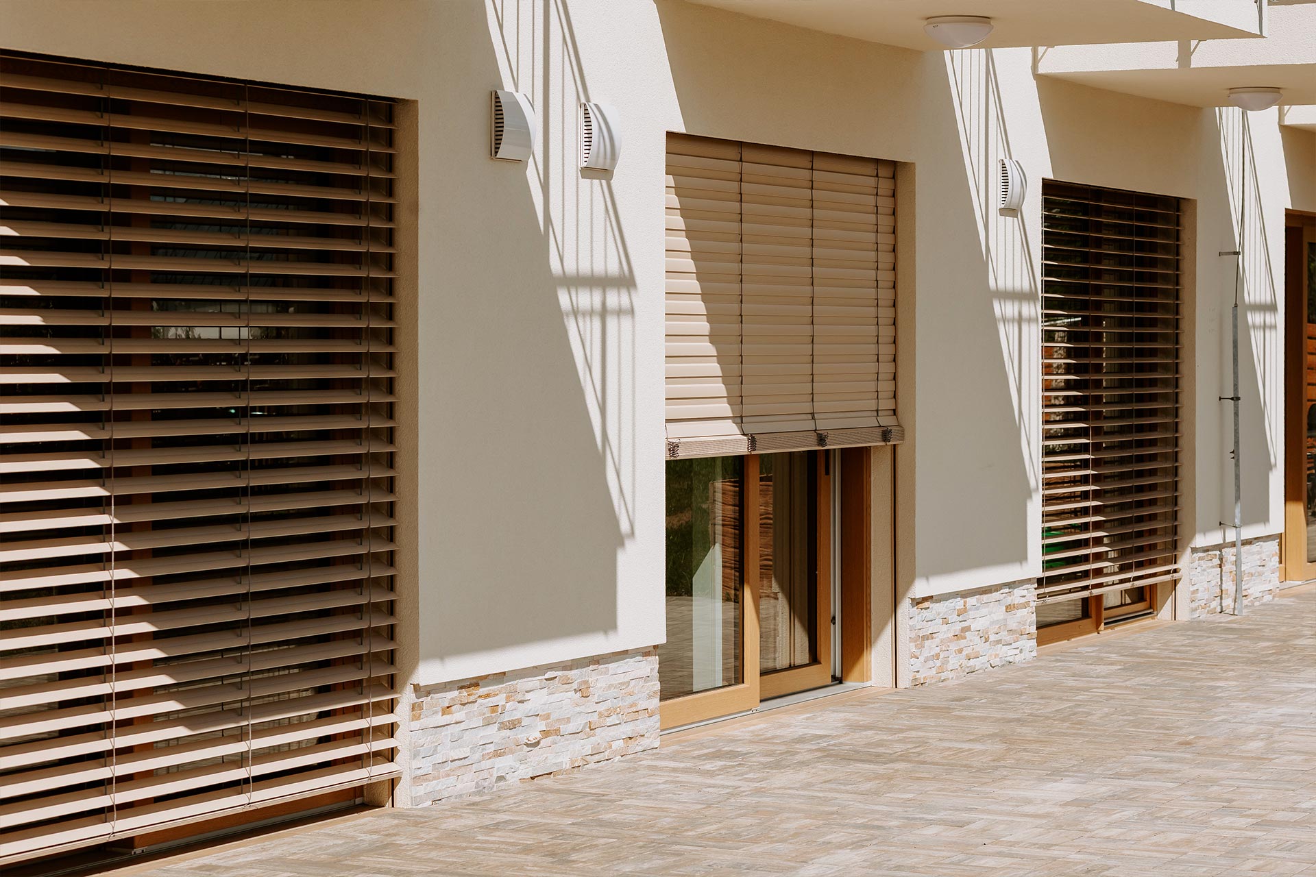 Exterior view of a modern beige building with large windows and horizontal wooden blinds, sunlight casting long shadows on the walls.