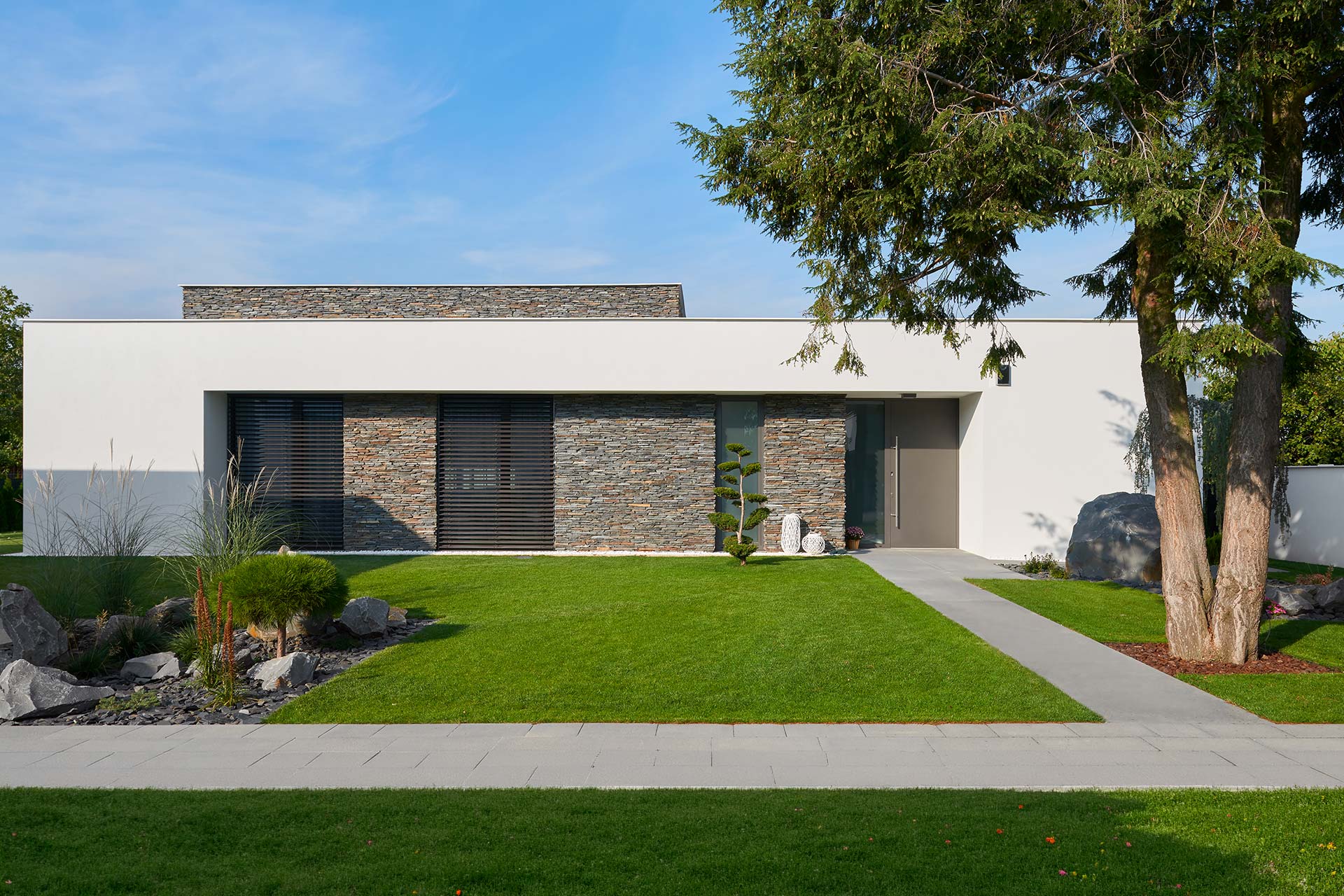 Front view of a modern, single-story home with a white facade, stone accents, and large windows set in a landscaped yard under a blue sky.